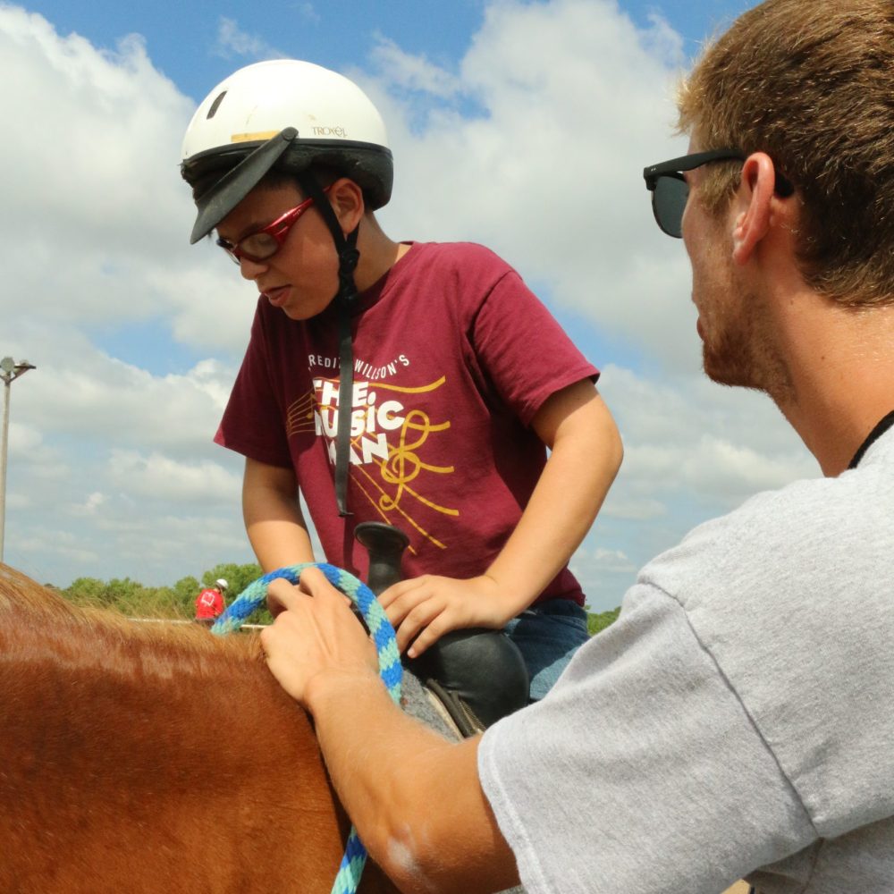 Horses, camp staff helping boy mount